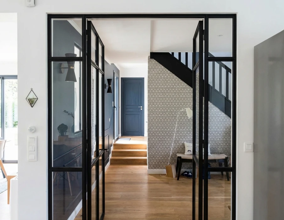 Couloir intérieur moderne avec verrière atelier à cadres métalliques noirs donnant sur un hall d'entrée